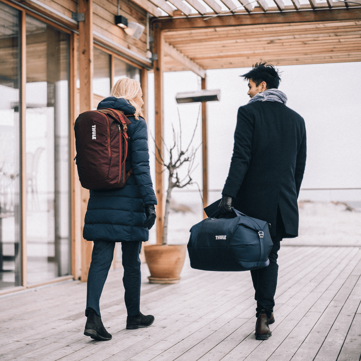 Two people walk down a wooden path by the beach with a Thule Subterra Weekender duffel bag.