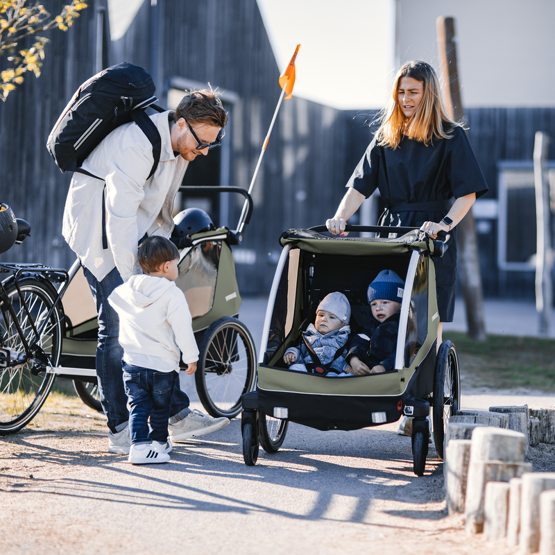 In a park, a father holds his kids' hand and a woman strolls with her kid in a Thule Courier child bike trailer.