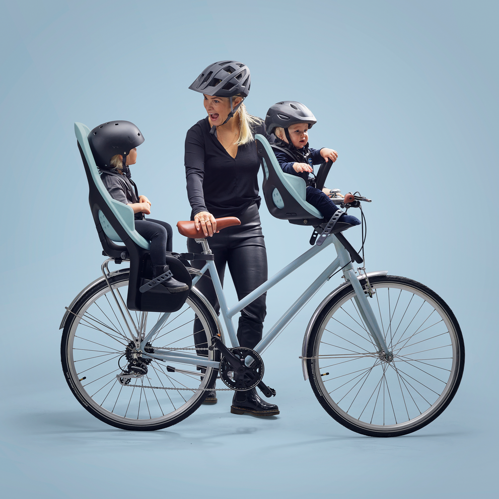 A woman stands with a blue bike, blue background and kids in a blue Thule Yepp 2 Maxi and Mini baby bike seat.