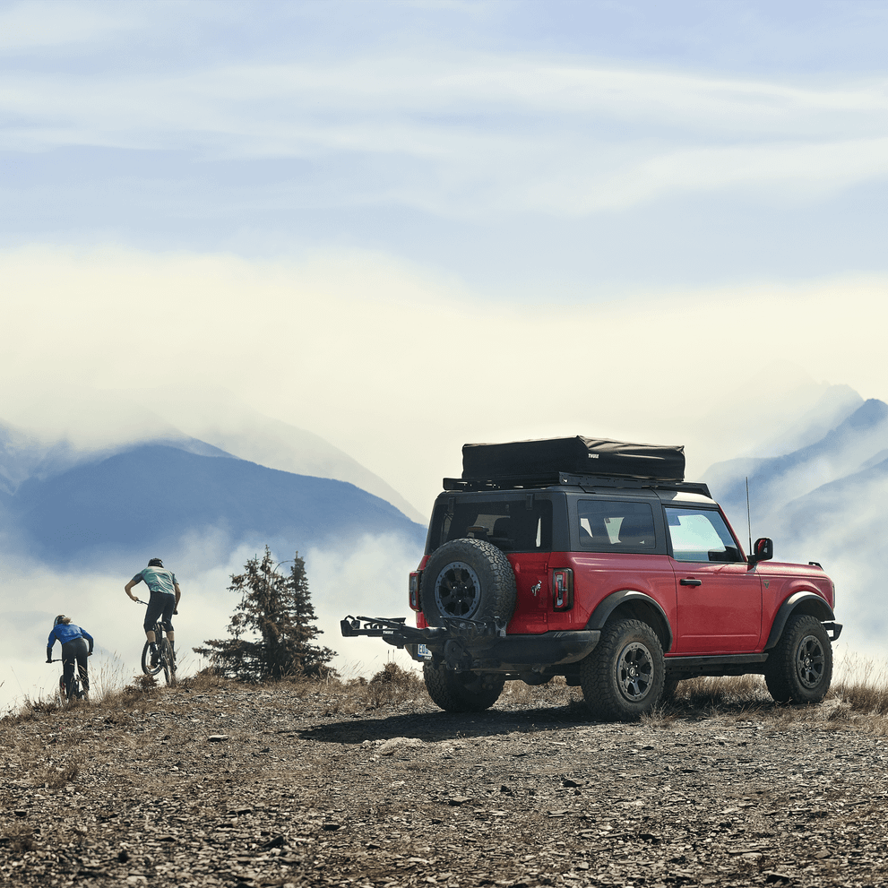 Parked in the mountains, cyclists cycle away from a red vehicle with a Thule T2 Pro XTR hitch bike rack.