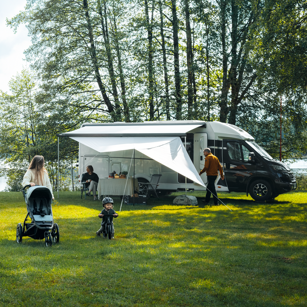 A motorhome with an extended Thule awning and panels setup on grass with a woman walking with her stroller and a child riding its bike.