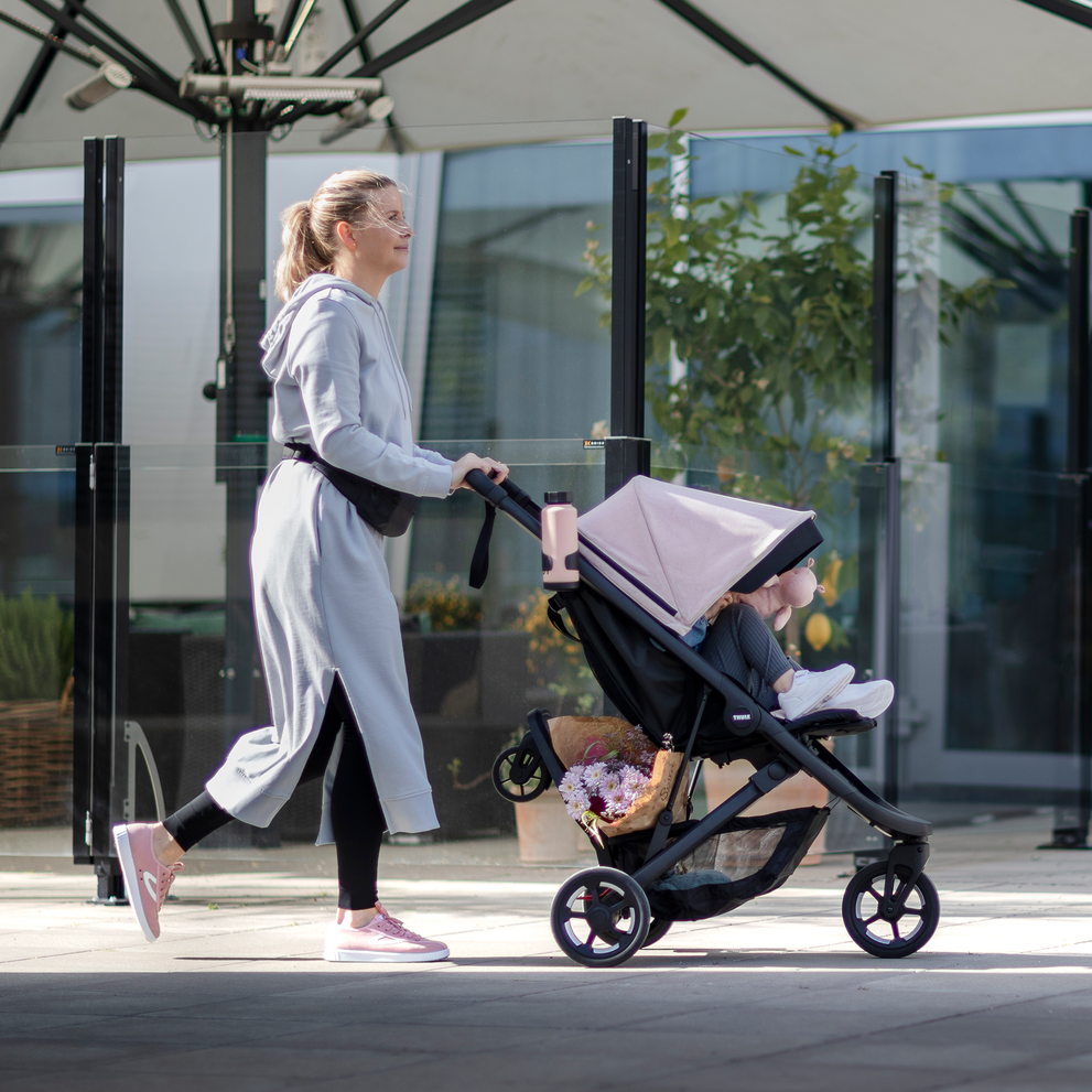 A woman walks down the street with her kid in a pink stroller and a Thule Rider Board.