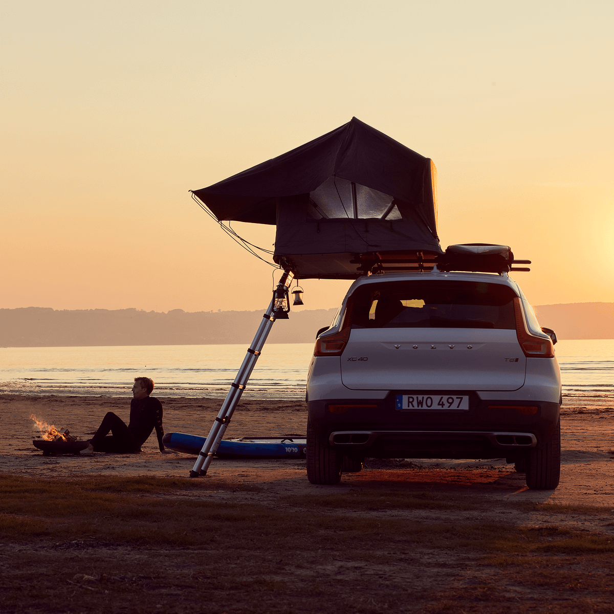 Two people are sitting by a fire on a beach next to a parked car with a roof top tent