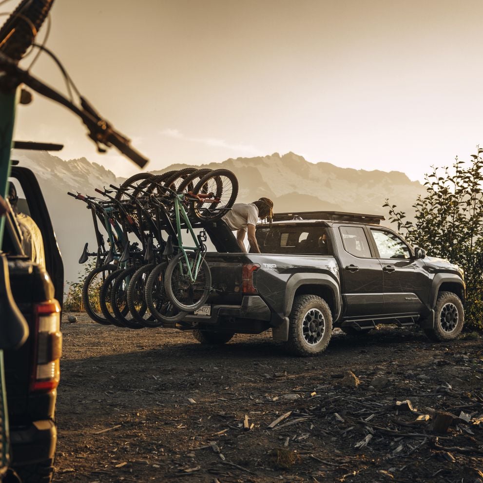A pickup truck with the verticle Thule ReVerft hitch bike rack fully loaded with mountain bikes, is pared in the mountains and a person is working in the cargo bed.