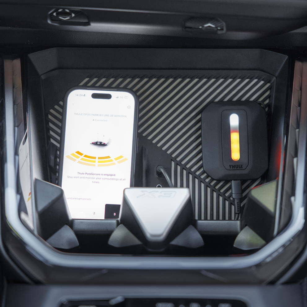 Interior view of a car showing the Thule Epos ParkSecure sensor unit and a smartphone displaying the app's parking assistance interface.