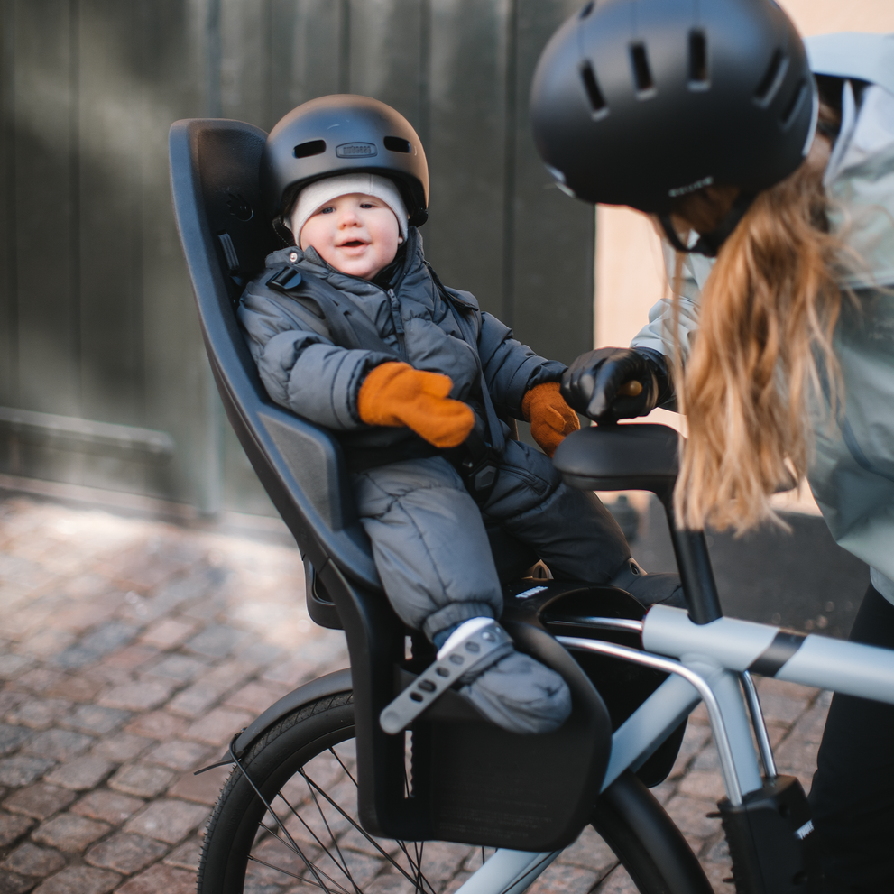 Next to a yellow wall, a woman speaks to her baby in a Thule Yepp 2 Maxi child bike seat.