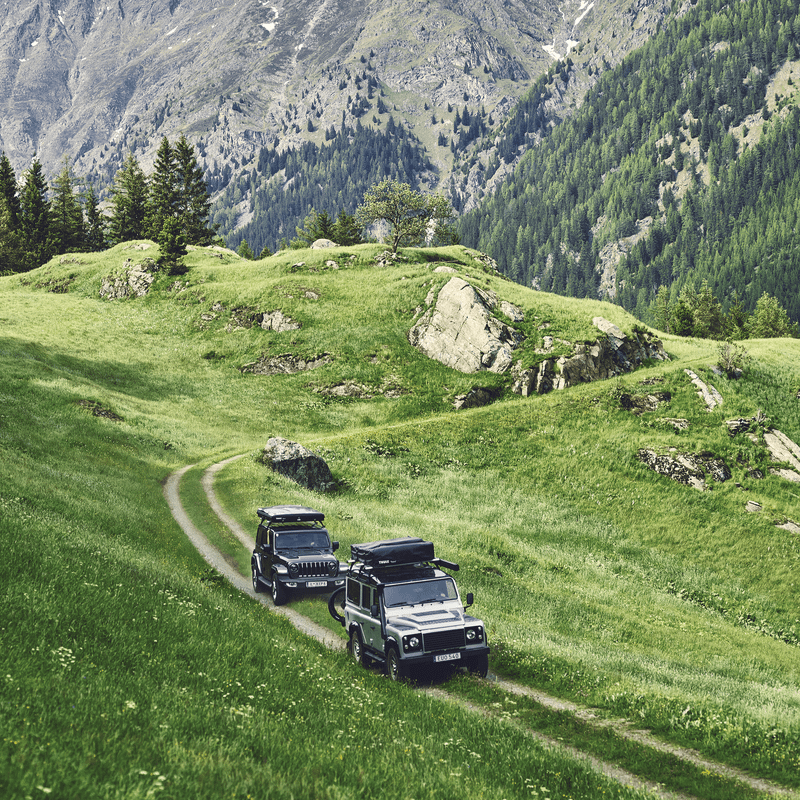 Two cars with roof top tents mounted are driving on a mountain road