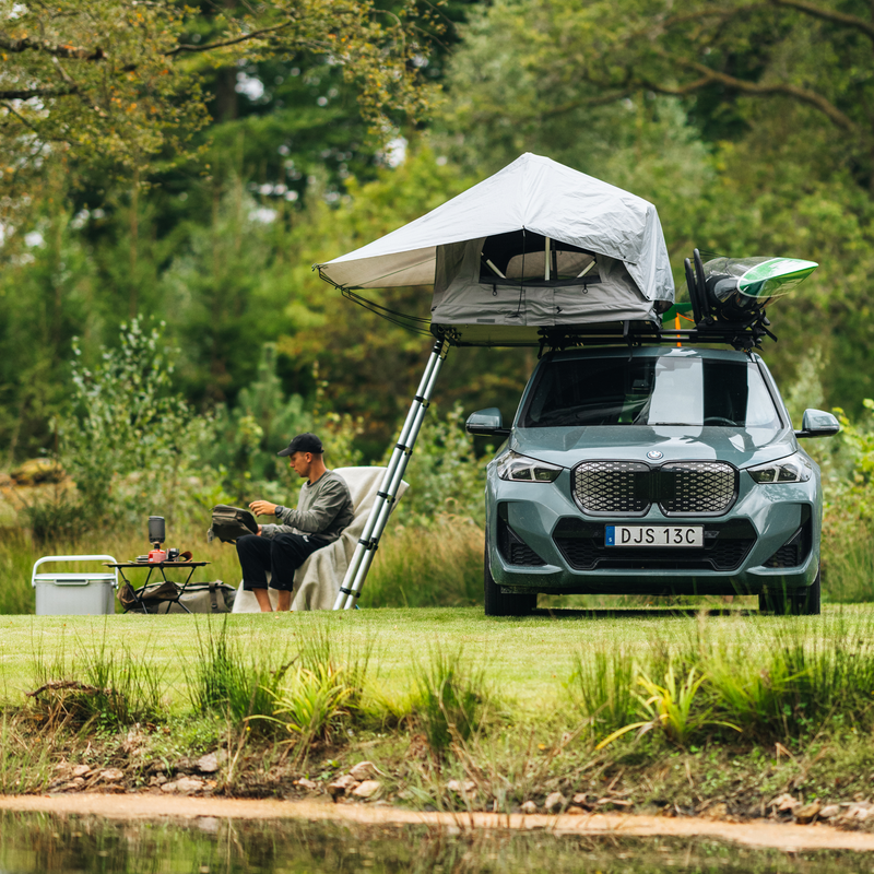 Thule Foothill 2 soft-shell rooftop tent unfolded and mounted on top of a car with a kayak, parked in an outdoor setting and a man sitting by his camp site.