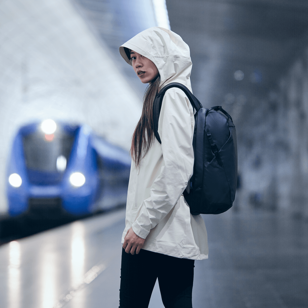 A woman with a white jacket and a hood waits at a train station, carrying a black Thule Tact backpack.