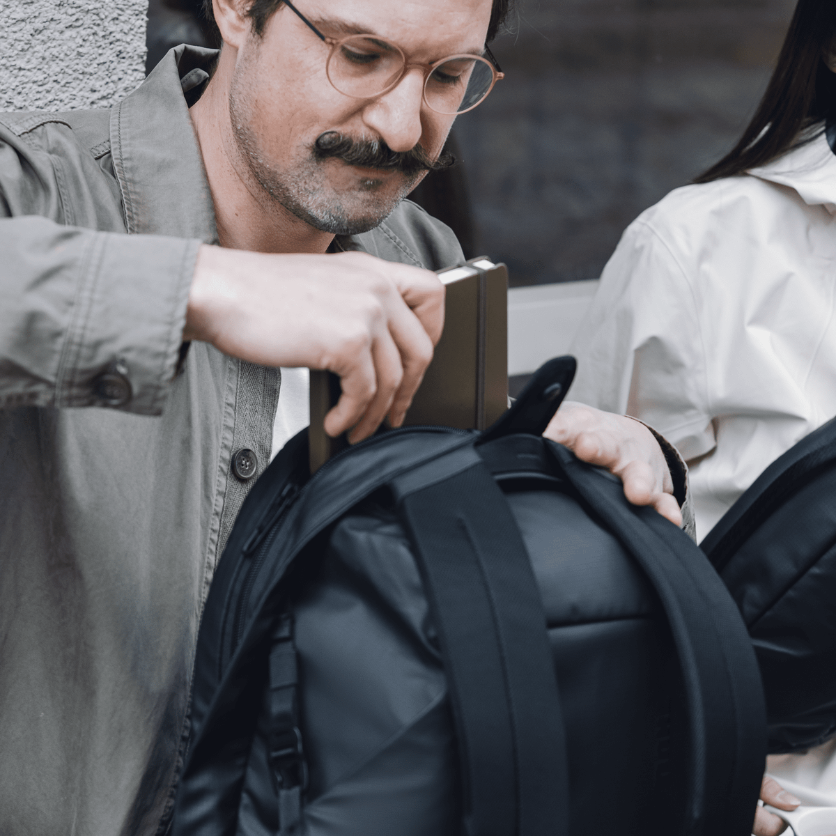 A man with glasses puts his notebook into a black Thule Tact backpack.