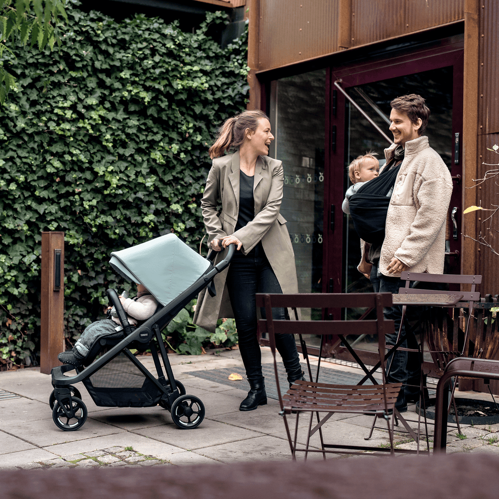 A woman with a blue Thule Shine compact stroller speaks to a man holding a baby in a carrier.
