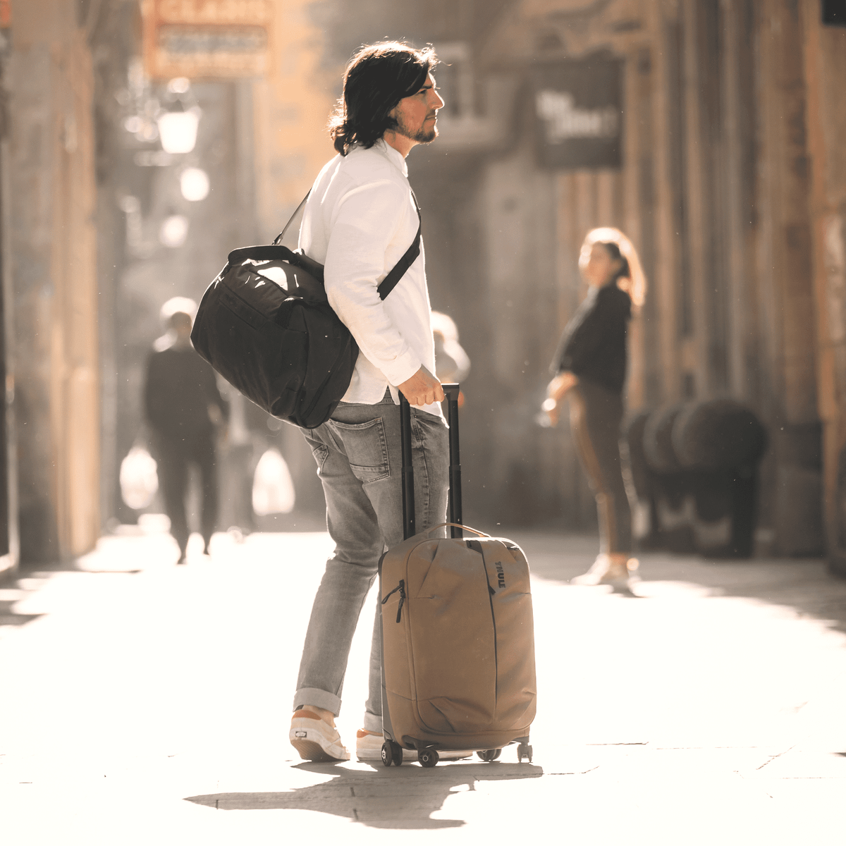 A man stands in the middle of a sunny street with a black duffel and tan Thule Aion Carry On Spinner suitcase.