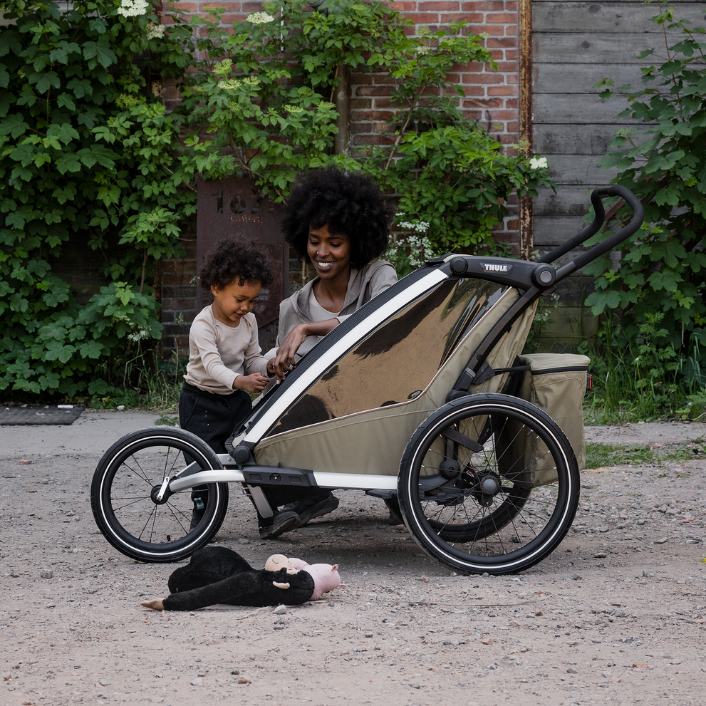 Parent with child beside a Thule Chariot bike trailer with all-terrain kit, parked outdoors on a gravel path.
