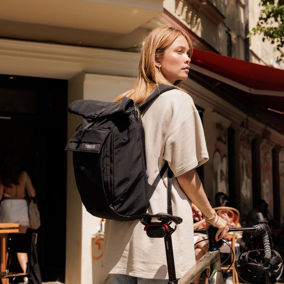 A woman stands on the street with her bike and a Thule Paramont backpack 24L.
