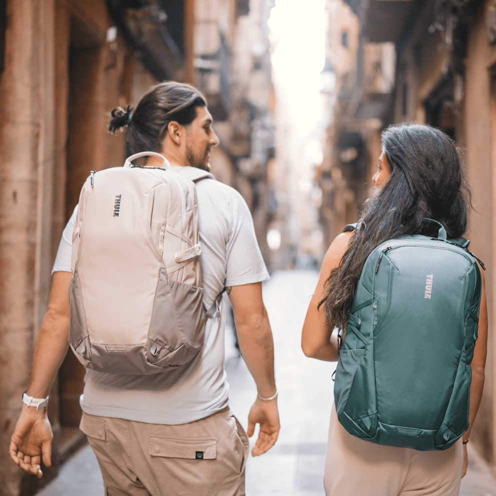 A man and women walk down a sunny, narrow city street with white and green Thule EnRoute backpacks.
