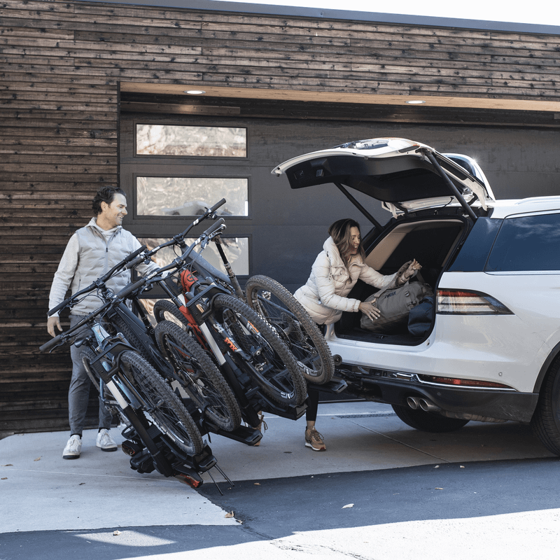 A man tilts the Thule Verse hitch bike rack so that a woman can access the trunk of a car.