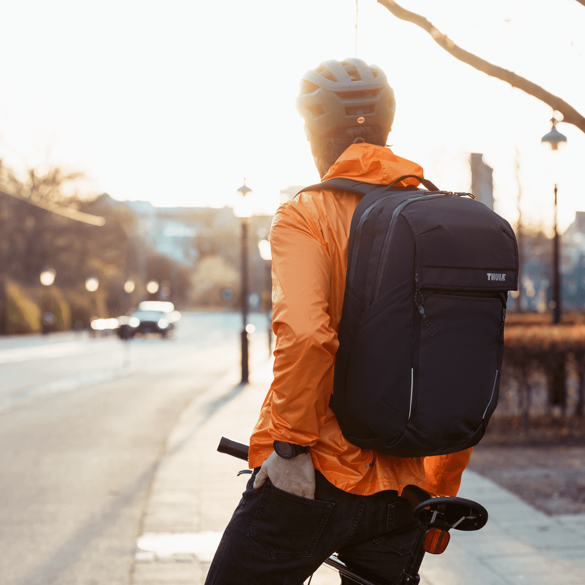 A cyclist by a city street looks at the sunshine, carrying a black 27L Thule Paramount Commuter Backpack.