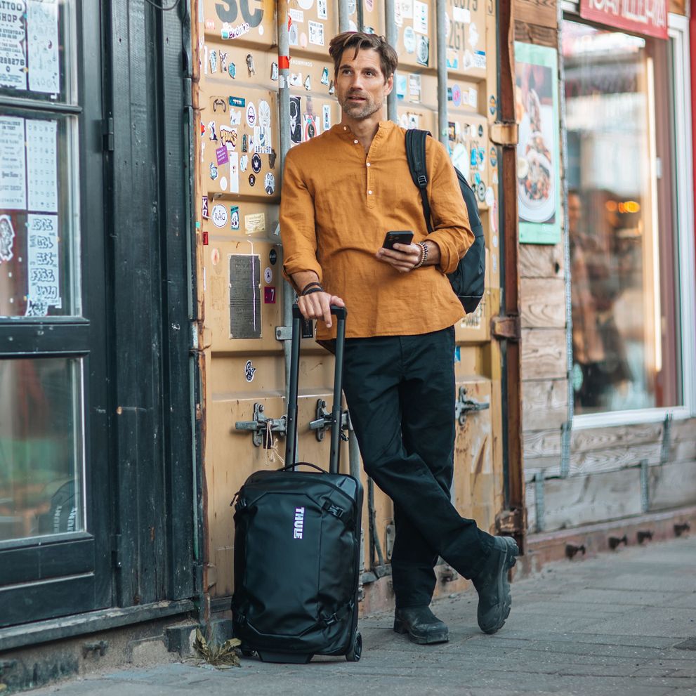 A man in an orange shirt is leaning against a wall with a black Thule Chasm carry-on suitcase.