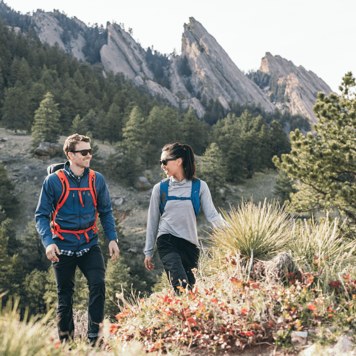 Two people hike in the mountains with Thule AllTrail 15L backpacks, wearing sunglasses.