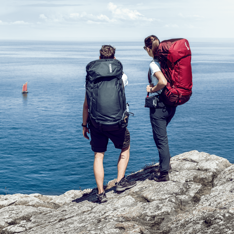 Two people stand on a rock next to the ocean carrying Thule GuidePost backpacks.