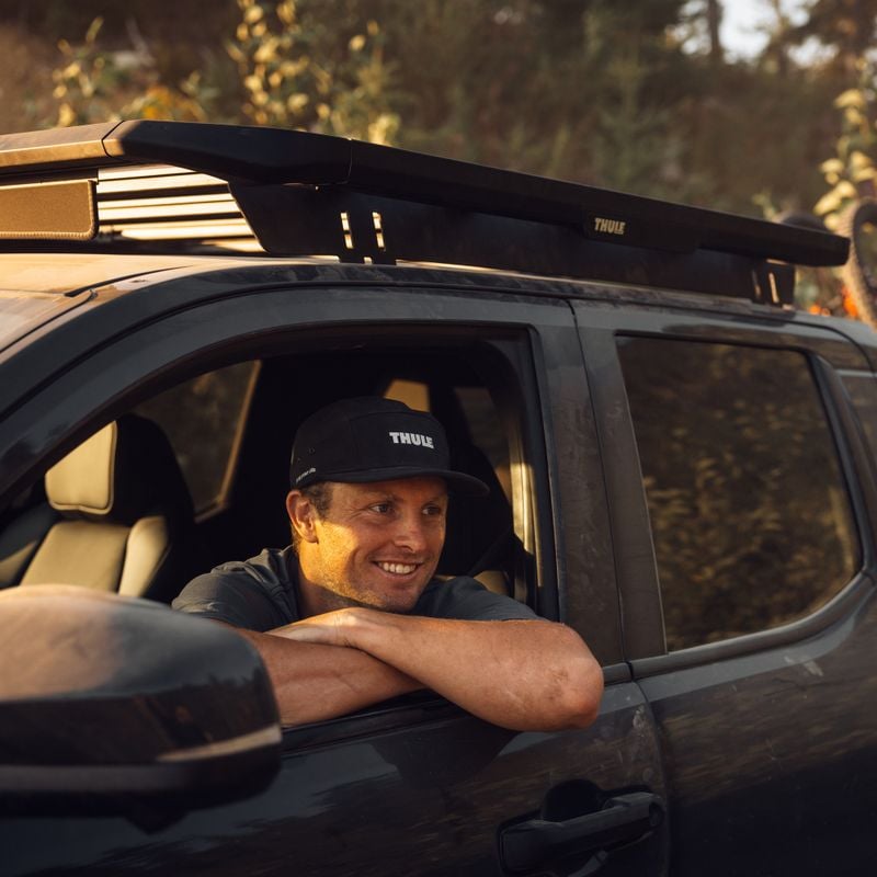 A man is sitting smiling in a off-road pickup truck with a Thule Caprock roof platform and a rail mount on the roof and a Thule ReVert hitch bike rack on the back of the truck with 6 bikes.