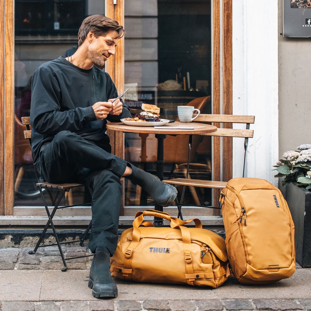 A man sits at a cafe with an orange Thule Chasm backpack and duffel.