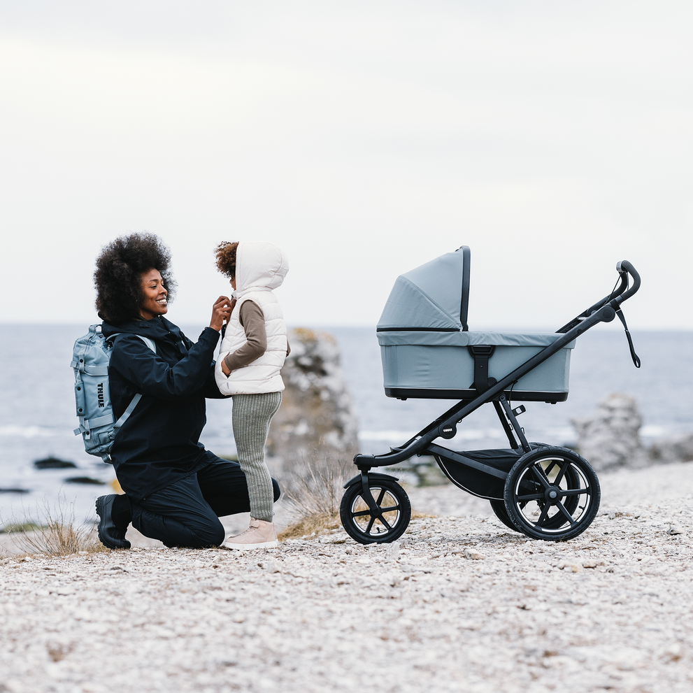 Pessoa empurrando Thule Urban Glide 3 com berço em caminho de cascalho, enquanto segura a mão de uma criança.