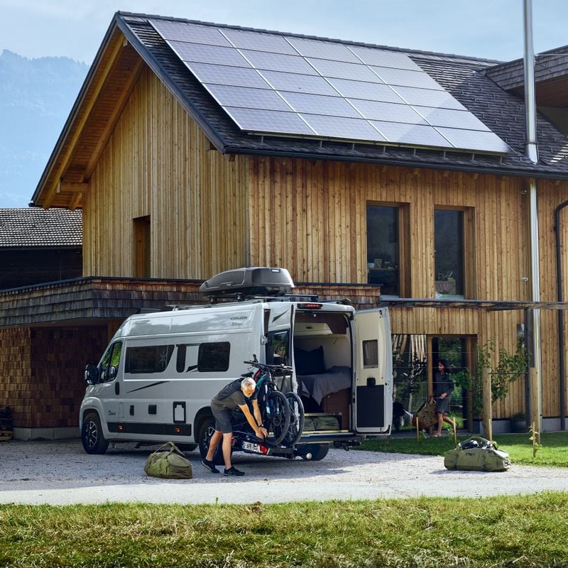 A white van is parked next to a woman building where the rear door is open with the Thule VeloSwing van ebike carrier and a bike rack attached.