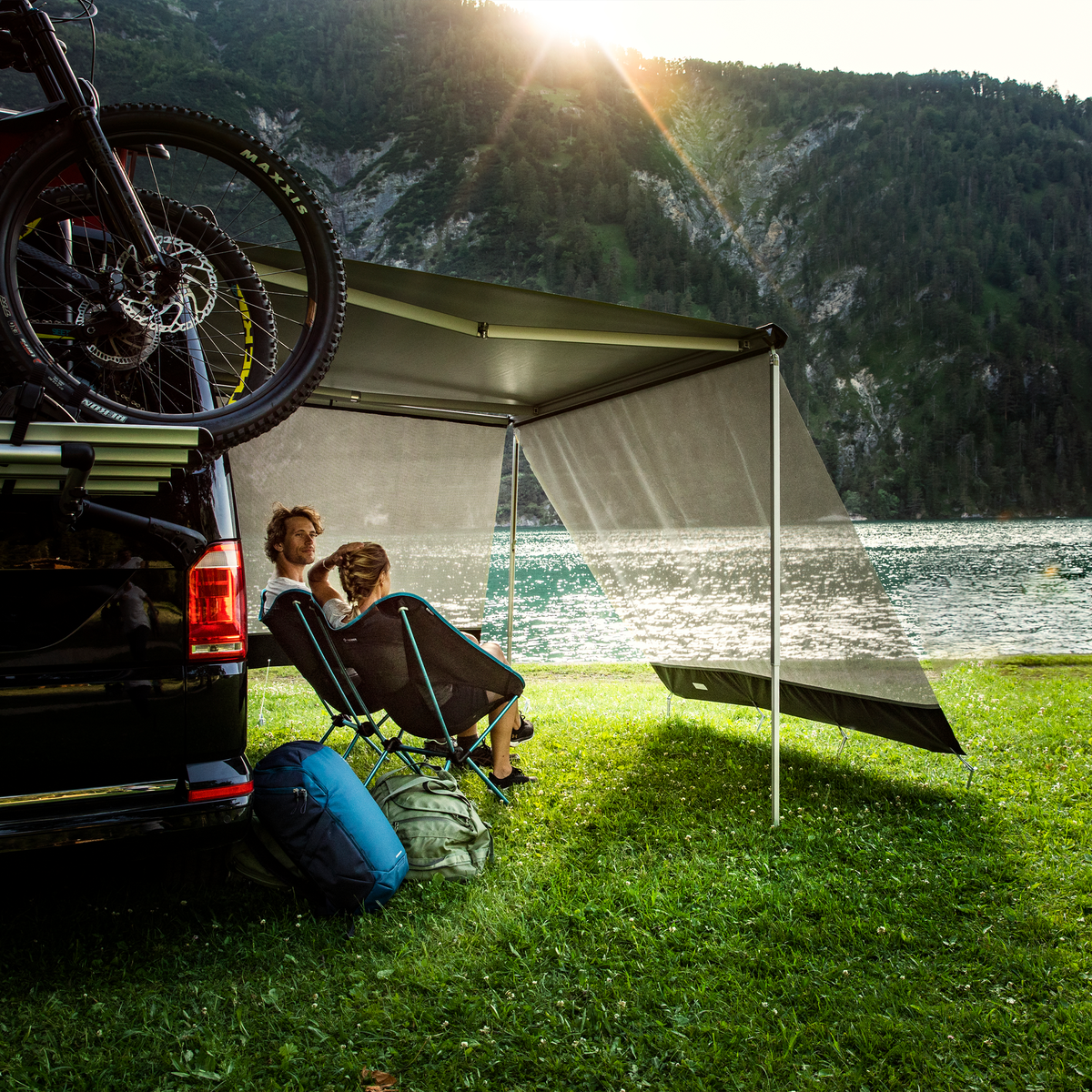 Two people sit by their black van with a Thule Sun Blocker Front awning side wall.