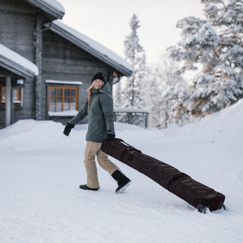 A woman stands in the snow outside a cottage wheeling her Thule RoundTrip travel ski bag.