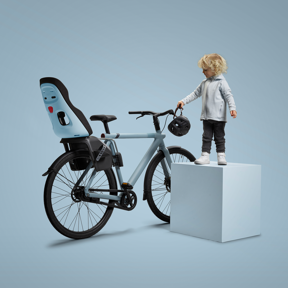 A child with curly blonde hair stands next to a modern grey bicycle, which is equipped with a blue and black child's bike seat mounted on the rear
