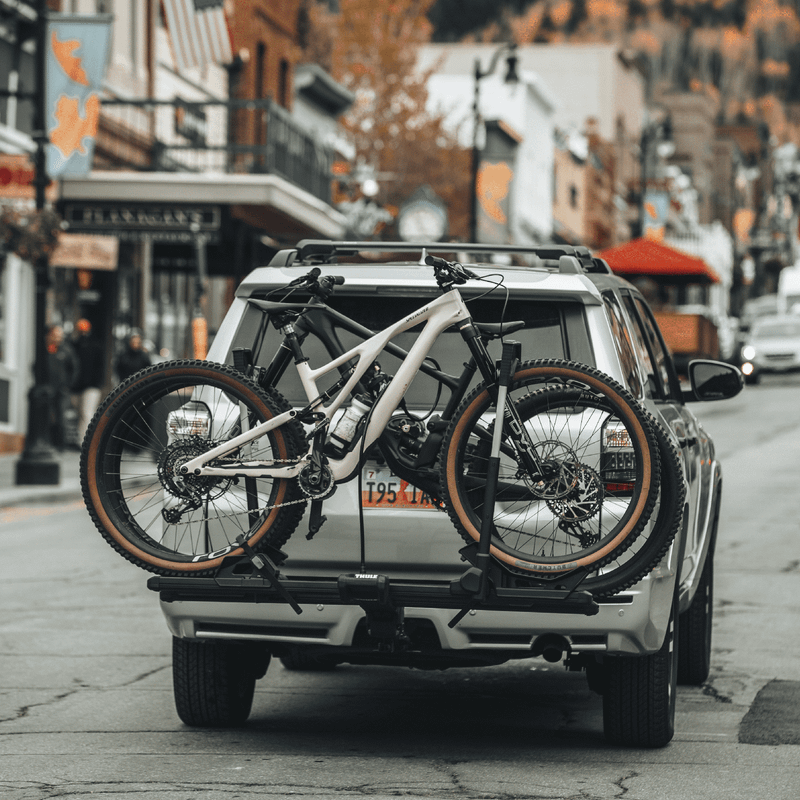 A silver SUV parked on a scenic downtown street with two mountain bikes securely mounted on a Thule Verse bike carrier.