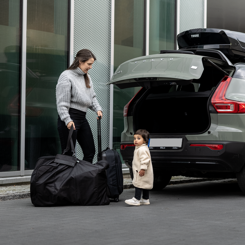 A woman stands next to the open trunk of her car with her kid and places a Thule Stroller Travel Bag  inside.