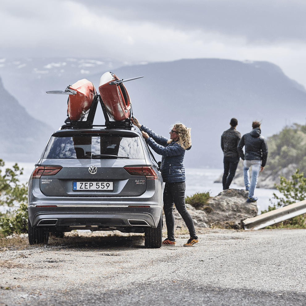 A woman is fastening a kayak on a car roof with a Thule Hull-a-Port Aero with two men in the background.