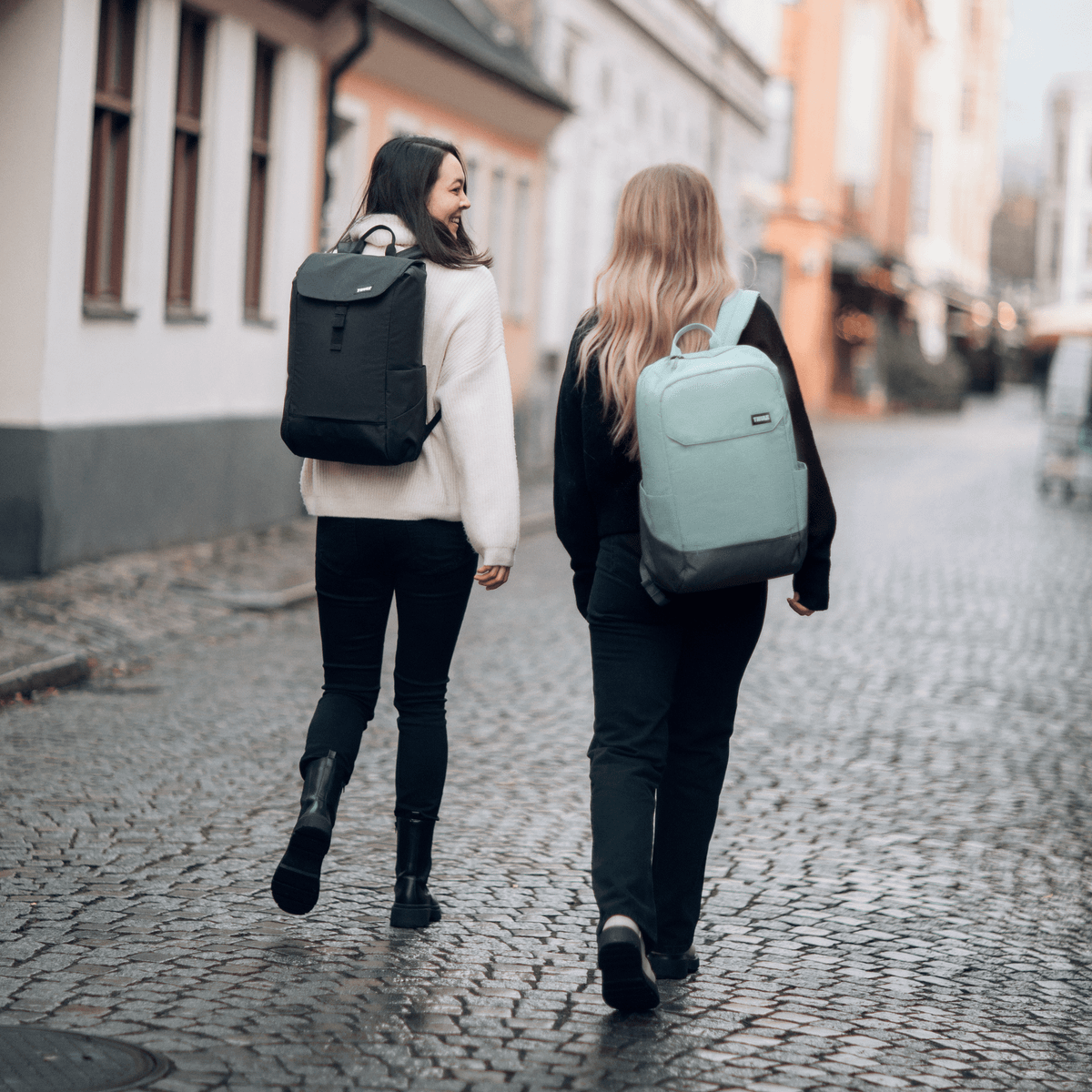 Two friends walk down a colorful, cobbled street carrying Thule Lithos backpacks.