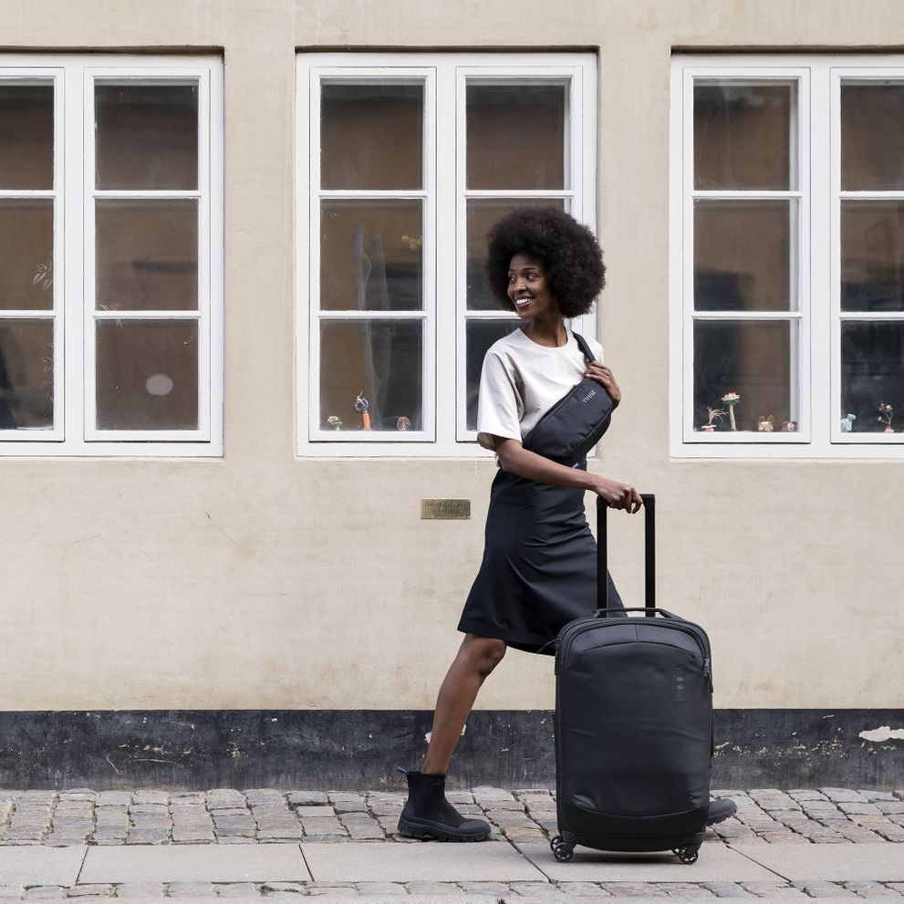 A woman walks down a cobbled street with a black Thule Subterra sling and Thule Subterra carry-on suitcase.