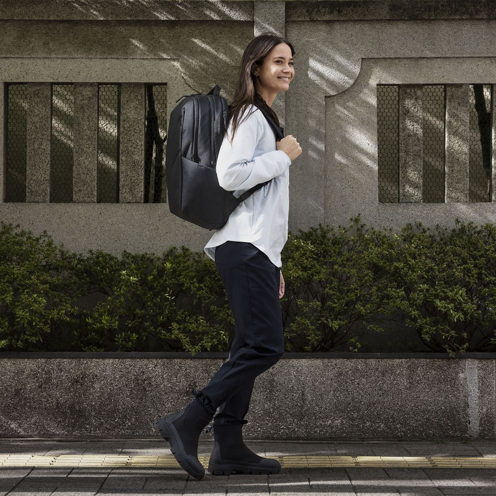 A woman walks down a sunny street while carrying a black Thule Subterra backpack.