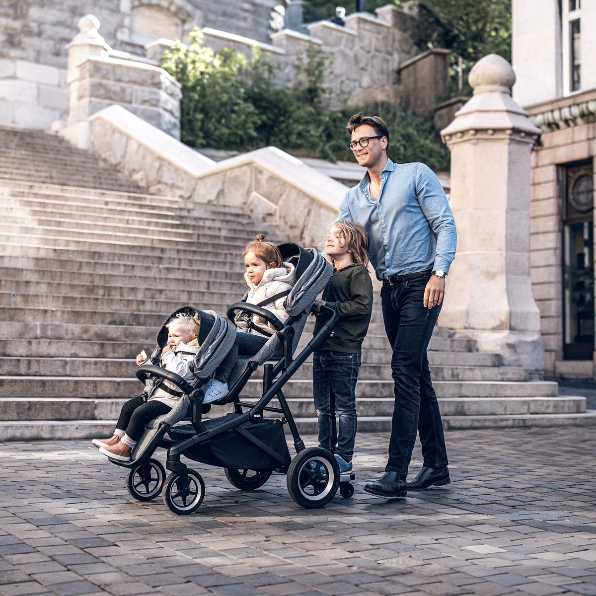 On a cobbled street next to stairs, a man pushes his two children in a Thule Sleek double stroller.
