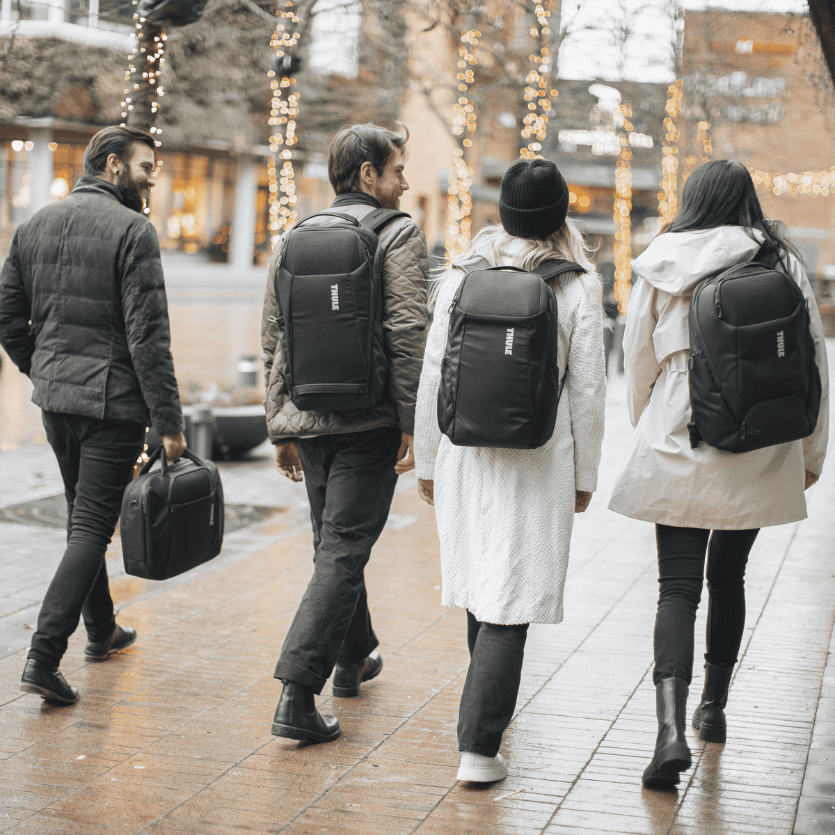 Four people walk down a rainy street wearing black Thule Accent backpacks.