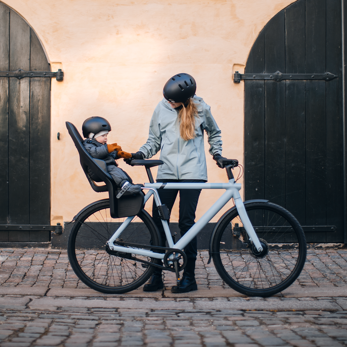 Next to a yellow wall, a woman speaks to her baby in a Thule Yepp 2 Maxi child bike seat.