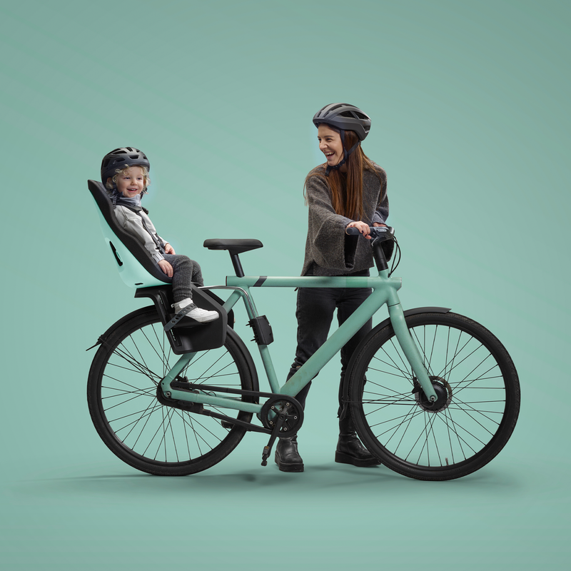 Un enfant joyeux portant un casque est assis dans un siège de vélo pour enfant monté à l'arrière d'un vélo vert pastel. Une femme, également avec un casque, se tient à califourchon sur le vélo, souriant à l'enfant