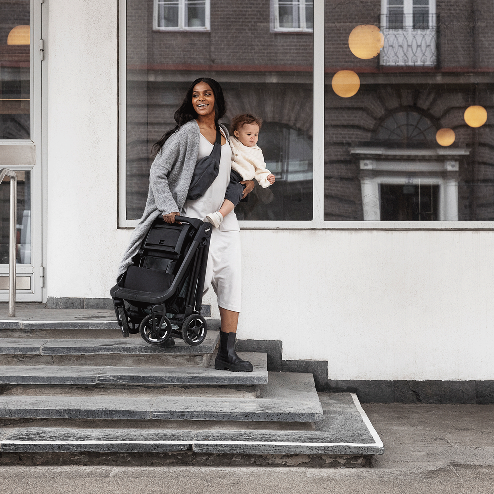 A woman walks down stairs holding her baby in one arm and her folded Thule Shine compact stroller in the other.