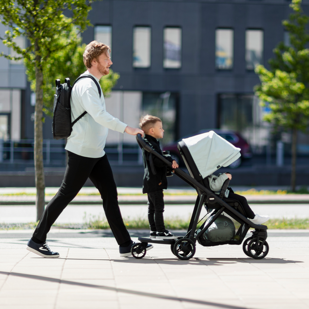 A man walks with one child in the blue stroller and another child on the Thule Rider Board.