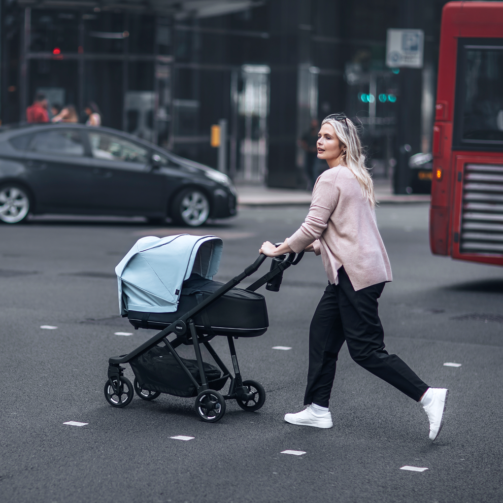 A woman walks across a crowded city street with a stroller that has a Thule Shine Air Purifier Canopy.