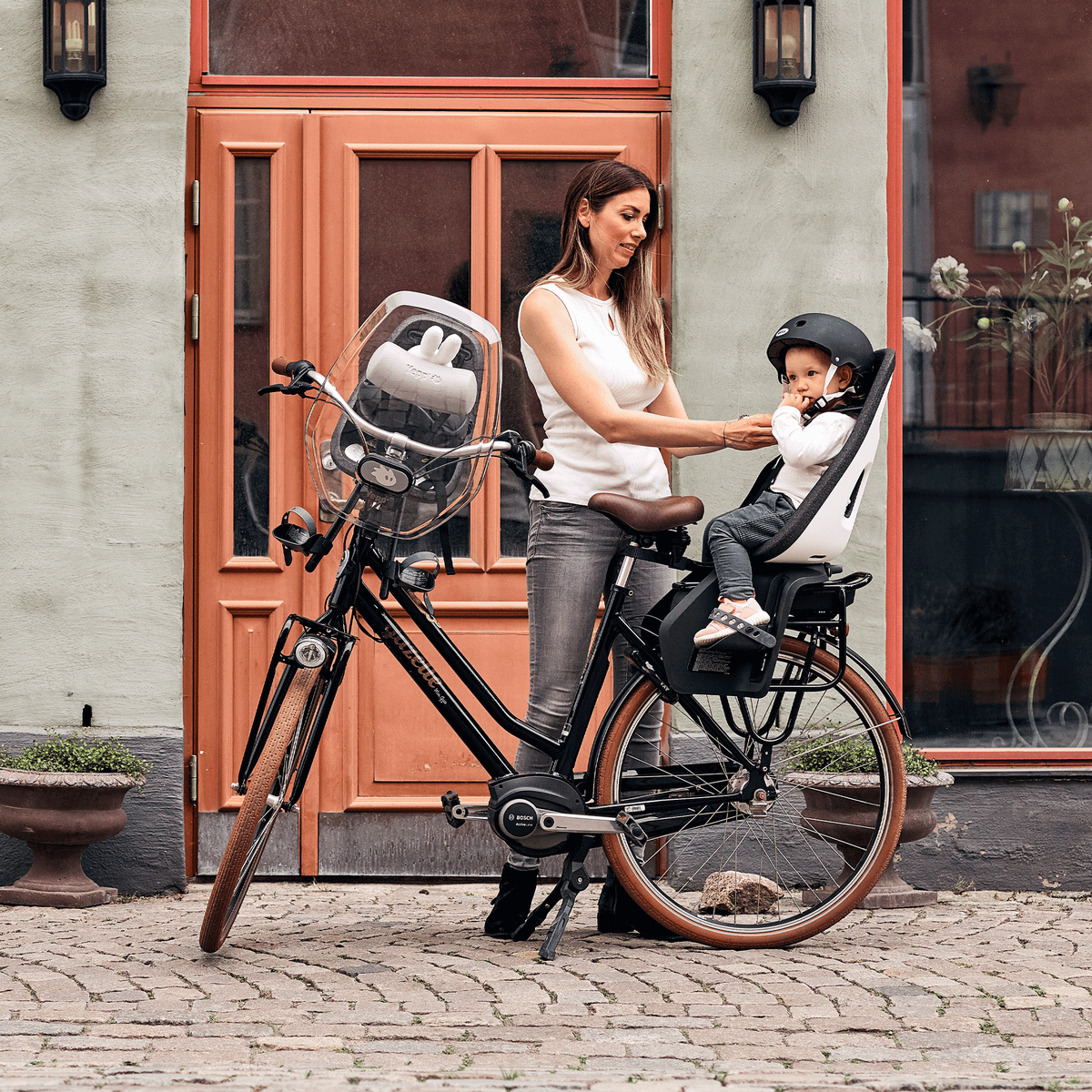 Outside a shop, woman adjusts the helmet of her child who sits in a Thule Yepp Nexxt child bike seat.