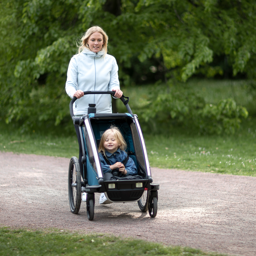 A woman in a blue jacket strolls with her child inside a blue Thule Chariot Cross kids' bike trailer.