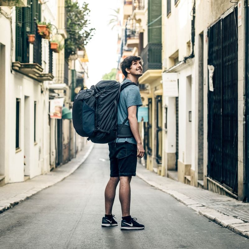 A man stands on an empty street looking up carrying a Thule Landmark 70L travel backpack.
