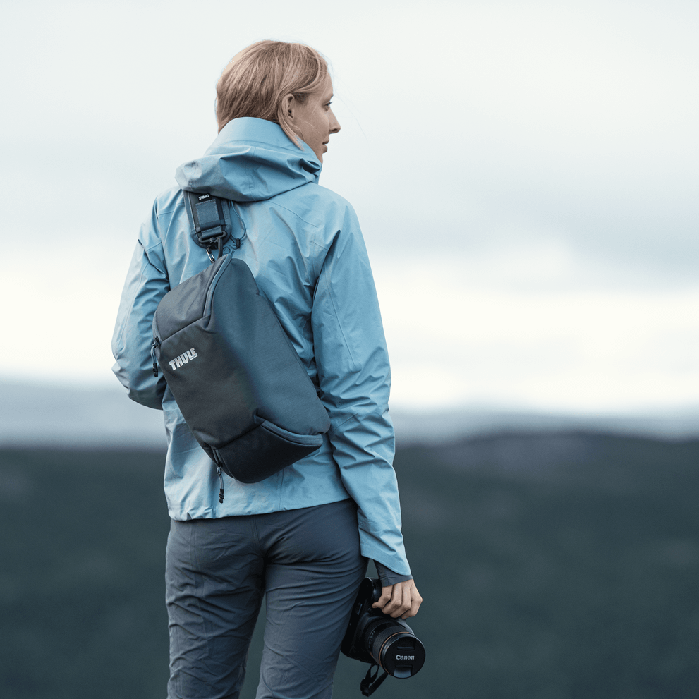 A woman holds  camera looking out into the distance carrying a blue Thule Covert DSLR camera backpack.