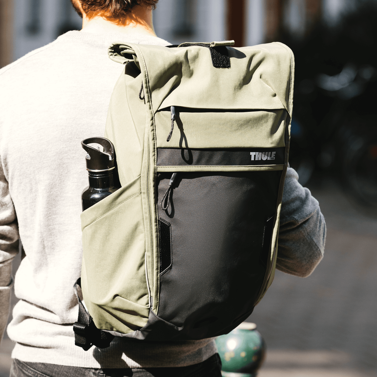 A man with his back tot he camera stands in the sunshine with a green 18L Thule Paramount Commuter Backpack.