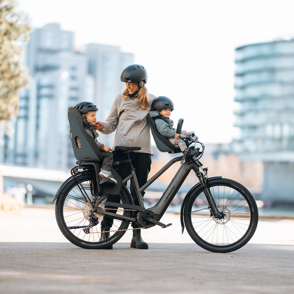 Adult standing beside a bicycle fitted with two Thule Yepp Nexxt 2 child bike seats, one in front and one in back, while interacting with two young children wearing helmets in an urban setting.
